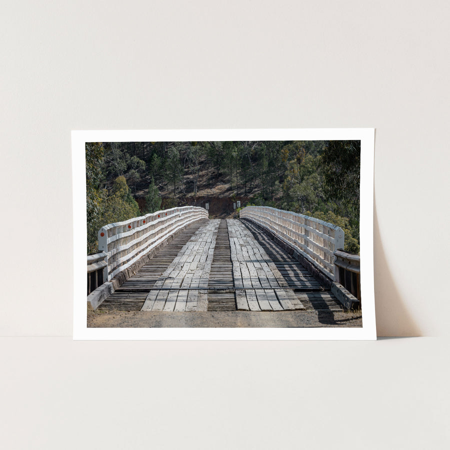 End-on view down the worn timber deck of McKillops Bridge in Deddick Valley, white handrails with red reflectors flanking the single lane as it runs toward a wooded hillside cutting.