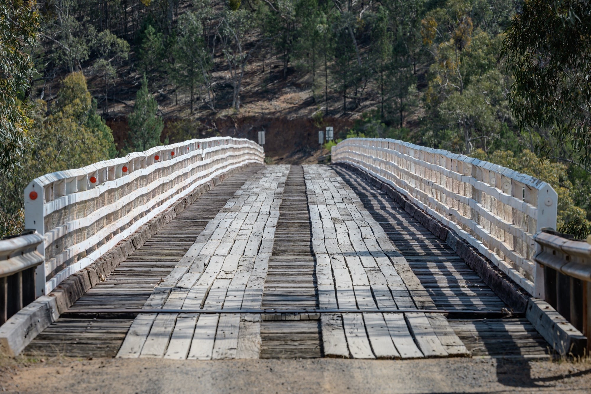 End-on view down the worn timber deck of McKillops Bridge in Deddick Valley, white handrails with red reflectors flanking the single lane as it runs toward a wooded hillside cutting.