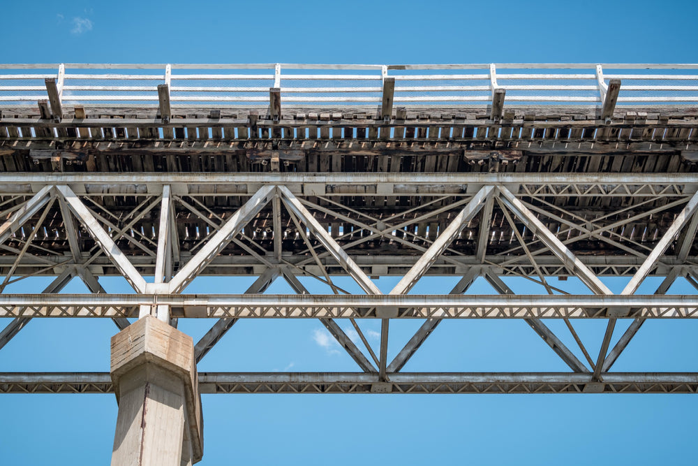 View from below McKillops Bridge in Deddick Valley, looking up at the electric-arc-welded steel Warren trusses and weathered timber decking against a clear sky, with a tall concrete pier at the left.