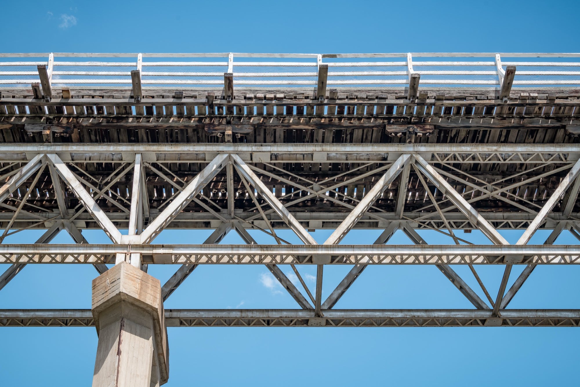 View from below McKillops Bridge in Deddick Valley, looking up at the electric-arc-welded steel Warren trusses and weathered timber decking against a clear sky, with a tall concrete pier at the left.