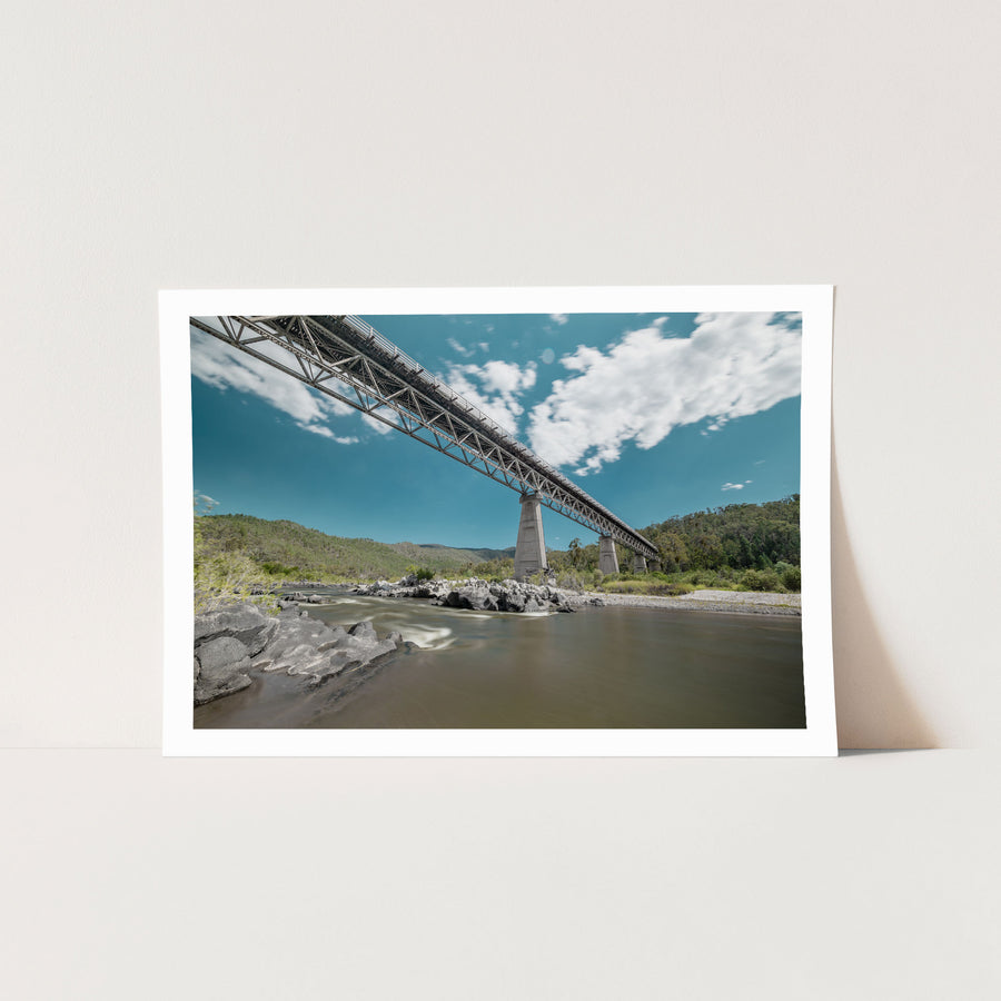 View from the rocky riverbed looking up at the steel Warren trusses and concrete A-frame pier of McKillops Bridge in the Deddick Valley, with the wide brown Snowy River running over exposed rock below and wooded hills behind.