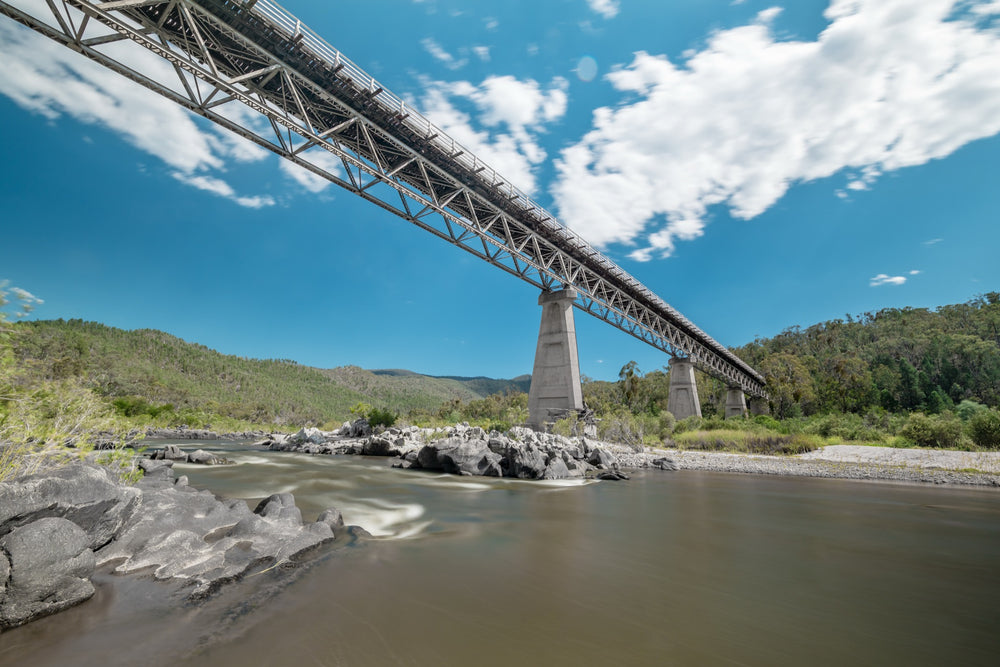 View from the rocky riverbed looking up at the steel Warren trusses and concrete A-frame pier of McKillops Bridge in the Deddick Valley, with the wide brown Snowy River running over exposed rock below and wooded hills behind.