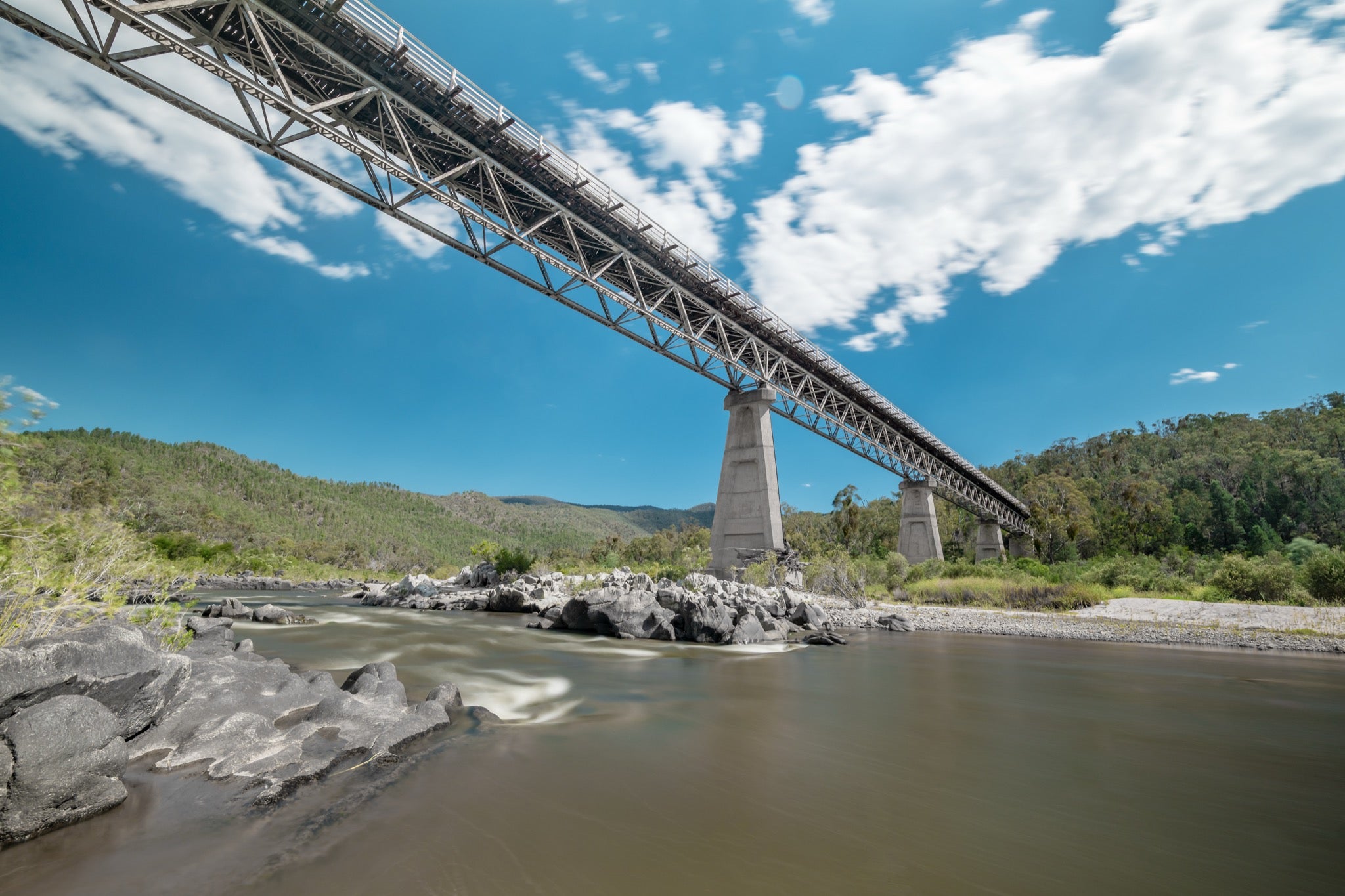 View from the rocky riverbed looking up at the steel Warren trusses and concrete A-frame pier of McKillops Bridge in the Deddick Valley, with the wide brown Snowy River running over exposed rock below and wooded hills behind.