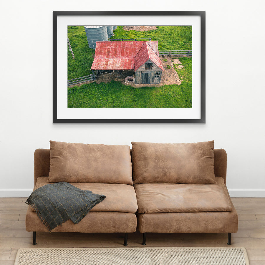 Aerial view of a timber cottage and shed under a faded red corrugated iron roof at Jembaicumbene, with a pig beside a stone trough near the doorway and two metal silos on the surrounding paddock.