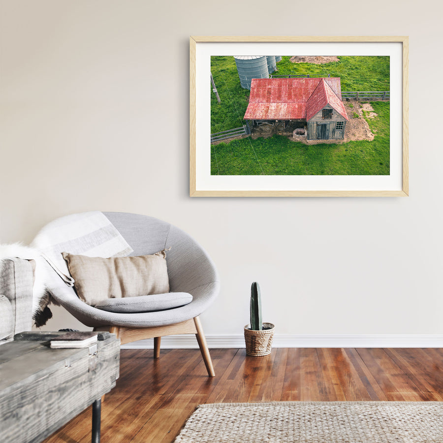 Aerial view of a timber cottage and shed under a faded red corrugated iron roof at Jembaicumbene, with a pig beside a stone trough near the doorway and two metal silos on the surrounding paddock.