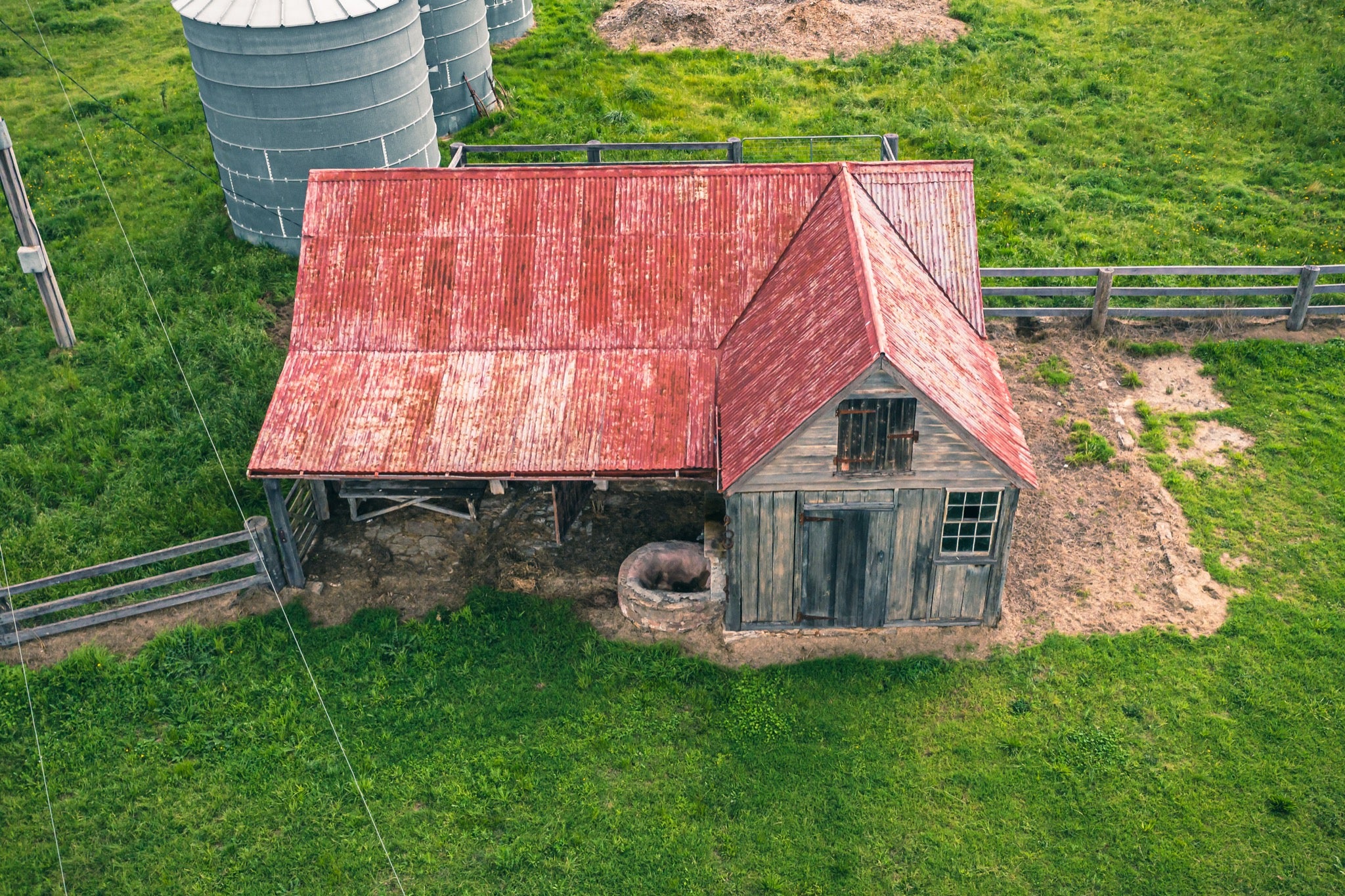 Aerial view of a timber cottage and shed under a faded red corrugated iron roof at Jembaicumbene, with a pig beside a stone trough near the doorway and two metal silos on the surrounding paddock.