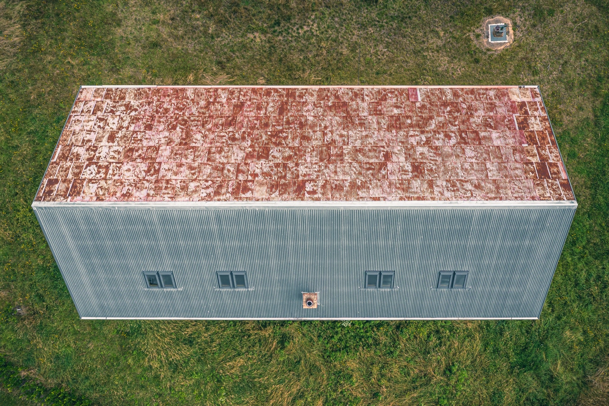Aerial view of a long corrugated iron farm building with a rust-mottled roof and small square windows, surrounded by mown grass at Jembaicumbene.