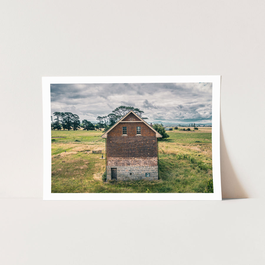 Gabled brick and stone end wall of the flour mill at Mill Pond Farm, Jembaicumbene, with two sash windows and a barred timber door at the granite base, surrounded by dry grass and a tall pine under an overcast sky.