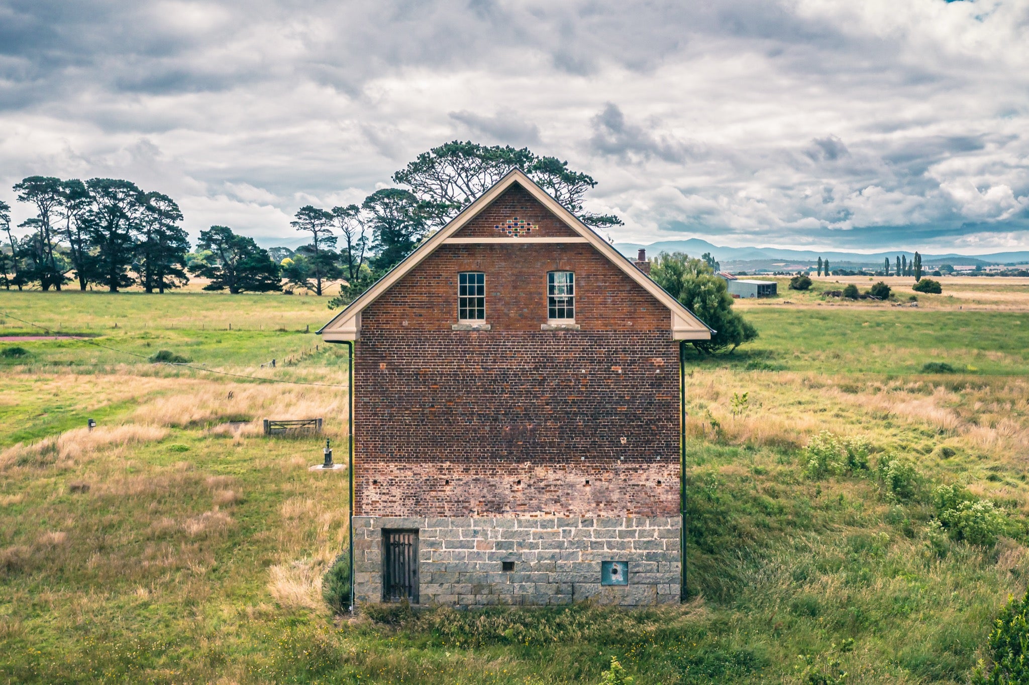 Gabled brick and stone end wall of the flour mill at Mill Pond Farm, Jembaicumbene, with two sash windows and a barred timber door at the granite base, surrounded by dry grass and a tall pine under an overcast sky.