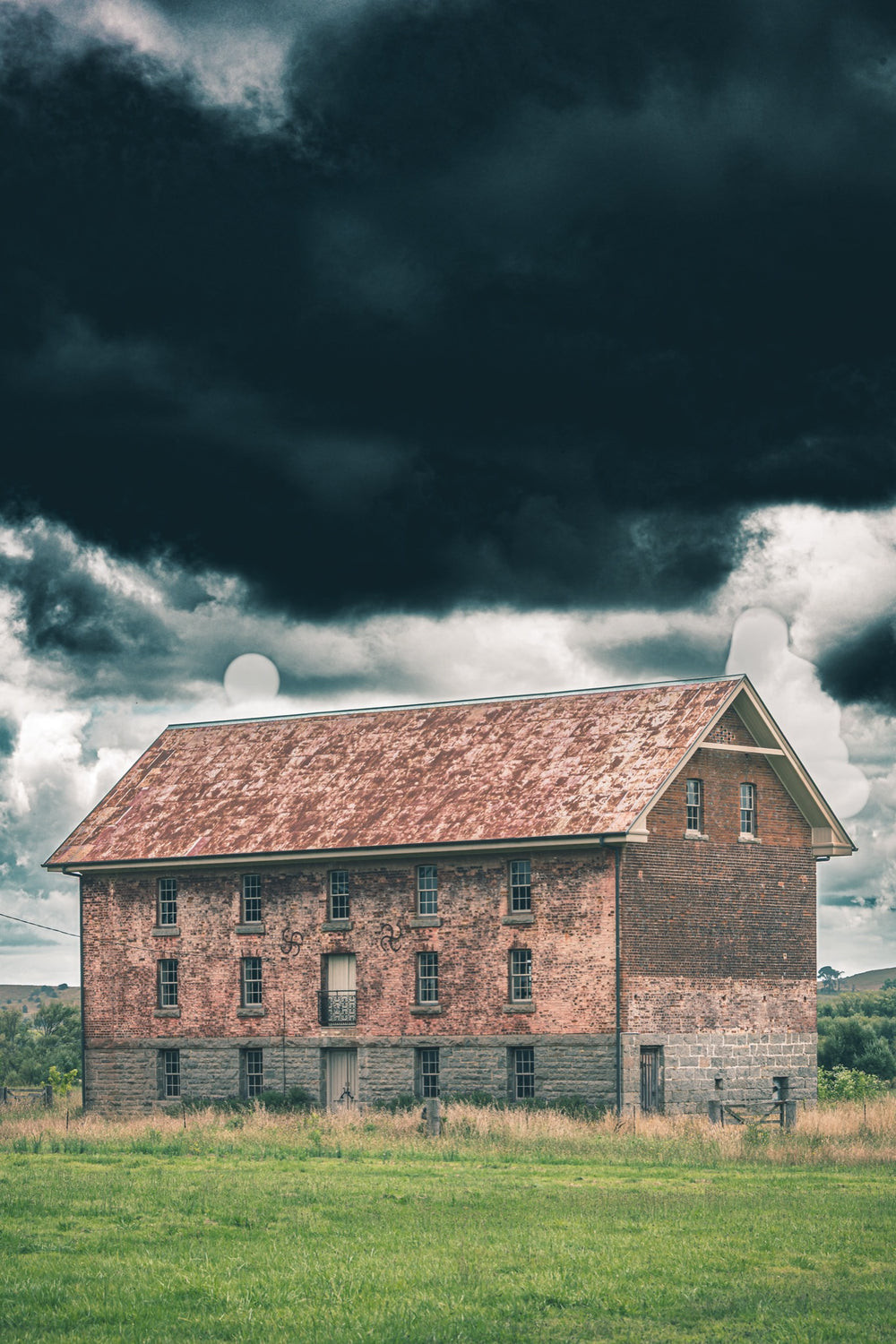 The four-storey brick and granite flour mill at Jembaicumbene, its multi-paned windows and central iron balcony visible against a heavy grey sky, photographed in open grassland near Braidwood.