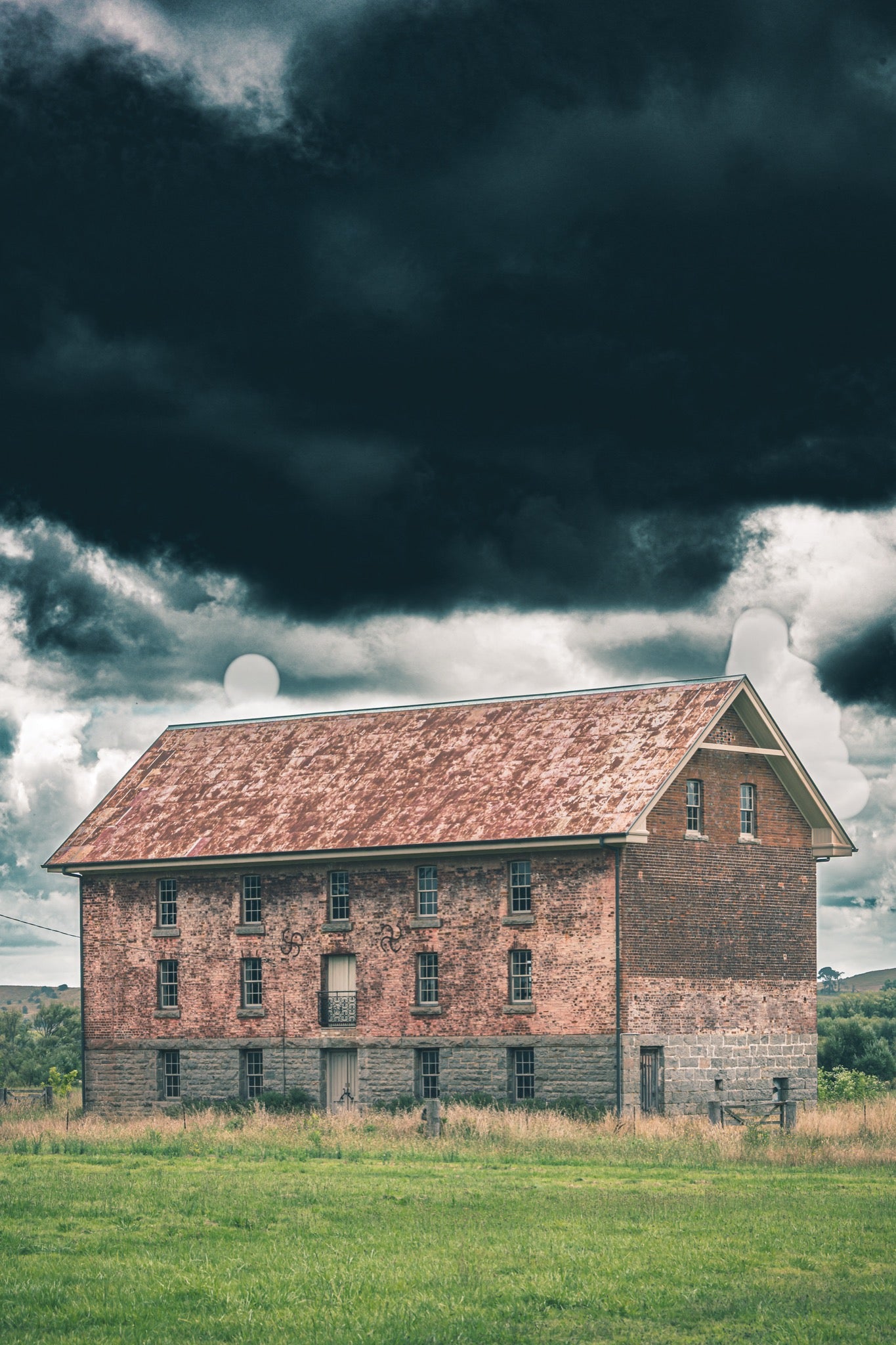 The four-storey brick and granite flour mill at Jembaicumbene, its multi-paned windows and central iron balcony visible against a heavy grey sky, photographed in open grassland near Braidwood.