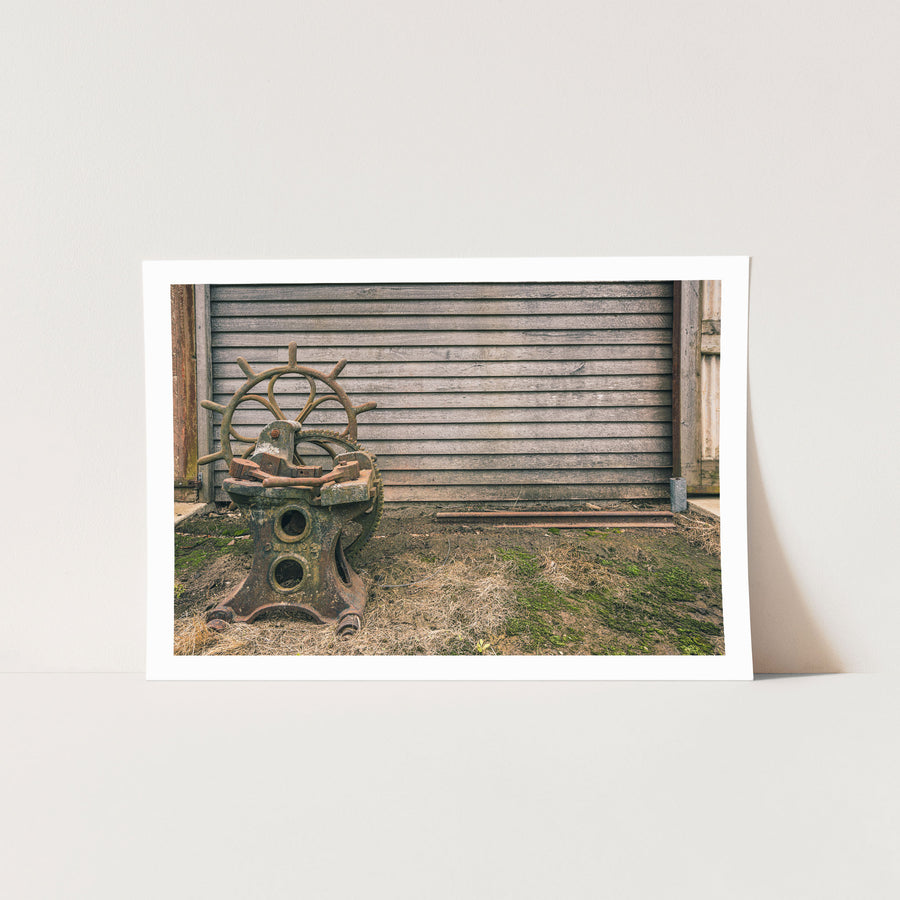 A rusted cast iron sharpening stand with a spoked hand wheel and toothed gear sits in dry grass against a weathered grey timber wall at Jembaicumbene.