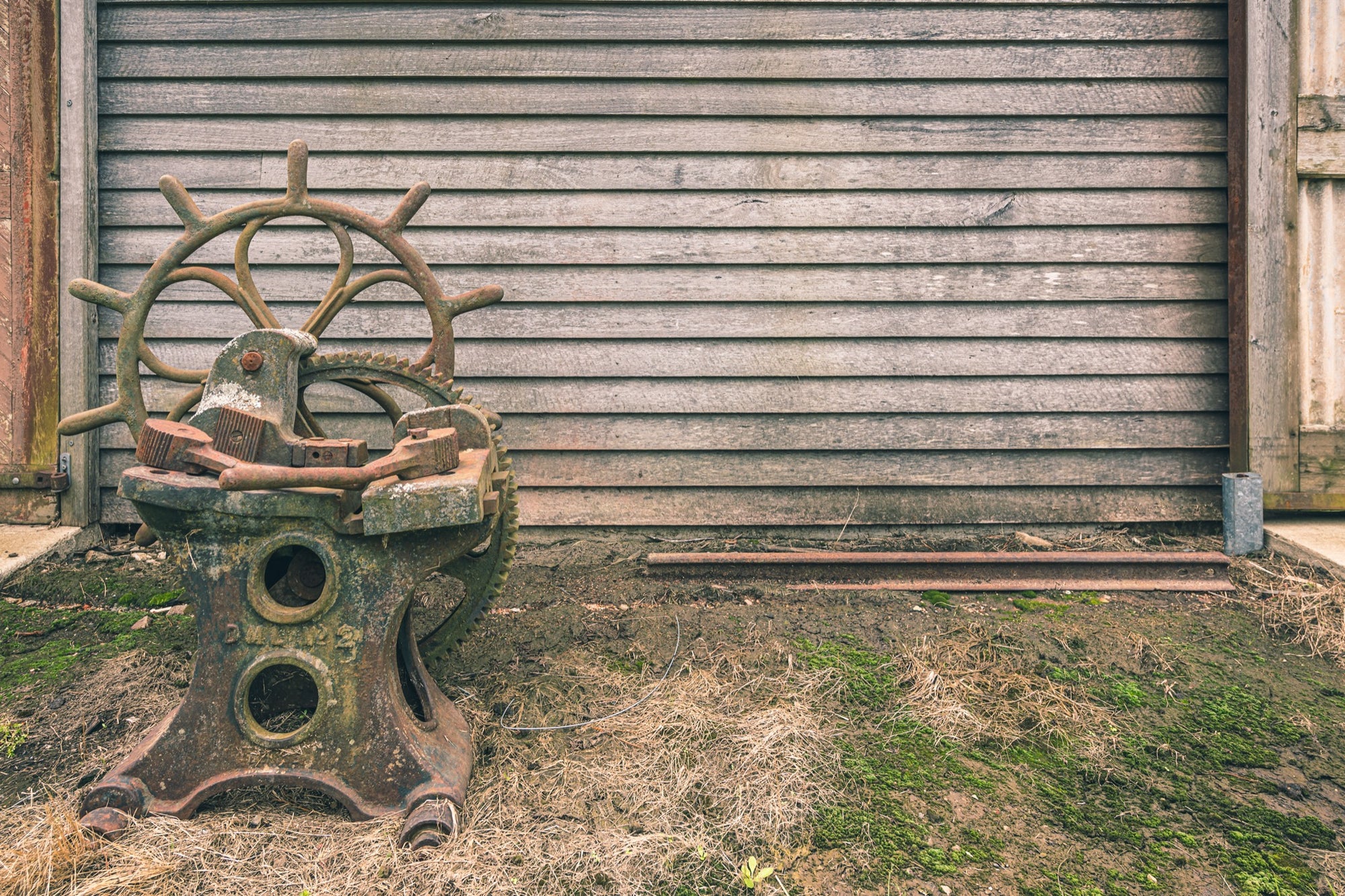 A rusted cast iron sharpening stand with a spoked hand wheel and toothed gear sits in dry grass against a weathered grey timber wall at Jembaicumbene.