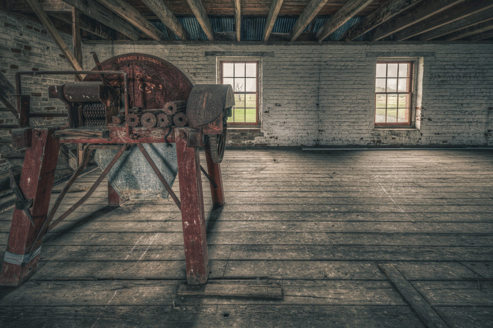 A red-painted chaff cutter with a large flywheel and toothed feed rollers on splayed timber legs, inside a whitewashed brick outbuilding at Mill Pond Farm, Jembaicumbene.