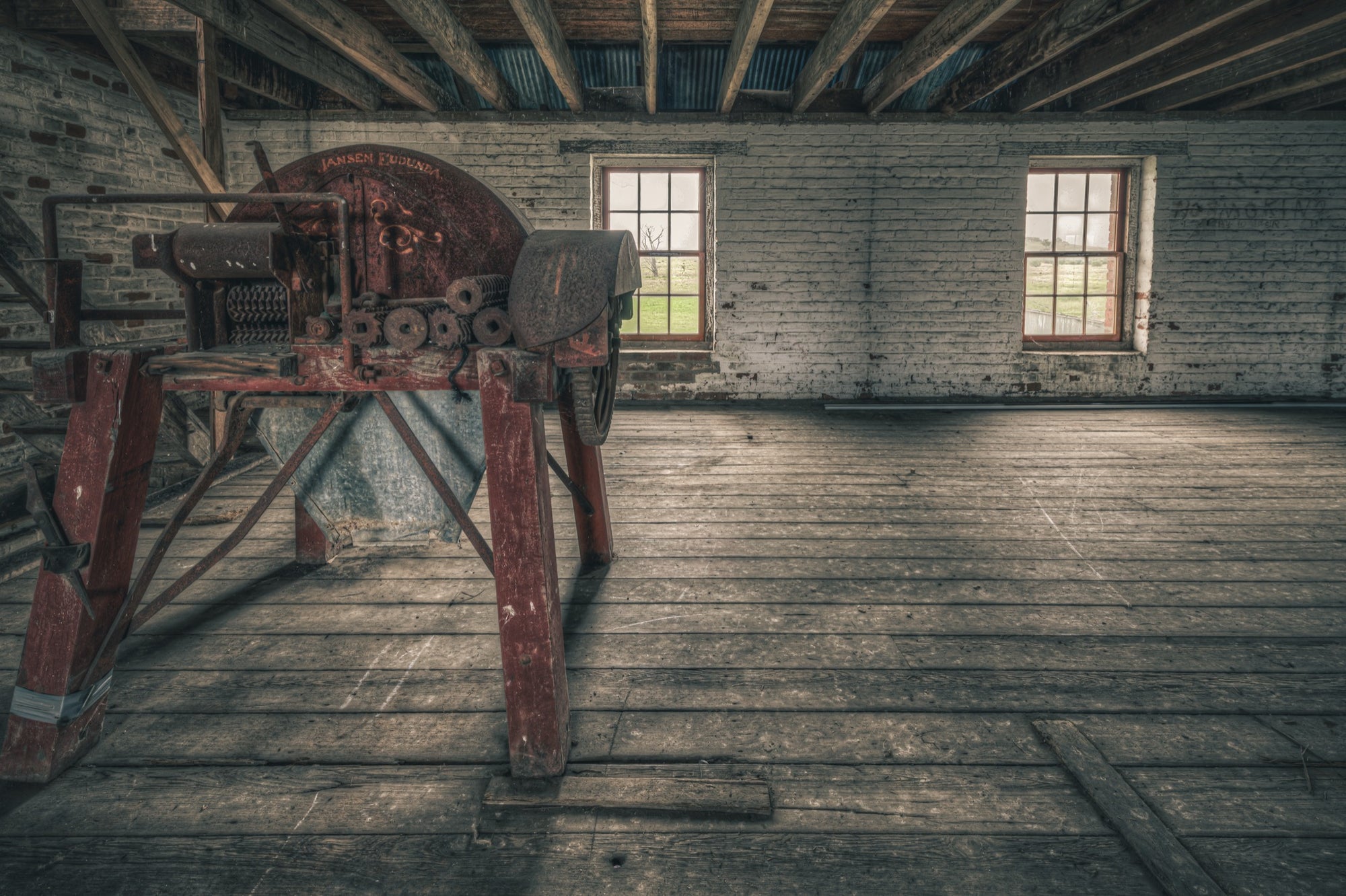 A red-painted chaff cutter with a large flywheel and toothed feed rollers on splayed timber legs, inside a whitewashed brick outbuilding at Mill Pond Farm, Jembaicumbene.