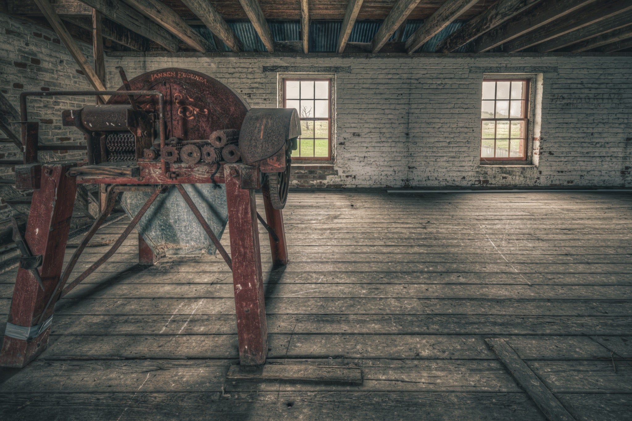 A red-painted chaff cutter with a large flywheel and toothed feed rollers on splayed timber legs, inside a whitewashed brick outbuilding at Mill Pond Farm, Jembaicumbene.