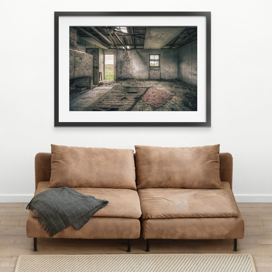 Interior of a derelict room at Jembaicumbene with a partly collapsed corrugated metal roof open to the sky, exposed timber rafters, rendered walls, a four-pane sash window, an open timber door to a paddock, and a torn red rug on broken floorboards.