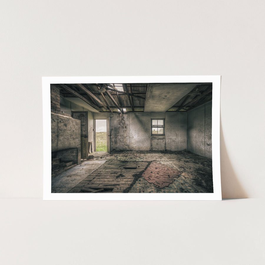 Interior of a derelict room at Jembaicumbene with a partly collapsed corrugated metal roof open to the sky, exposed timber rafters, rendered walls, a four-pane sash window, an open timber door to a paddock, and a torn red rug on broken floorboards.