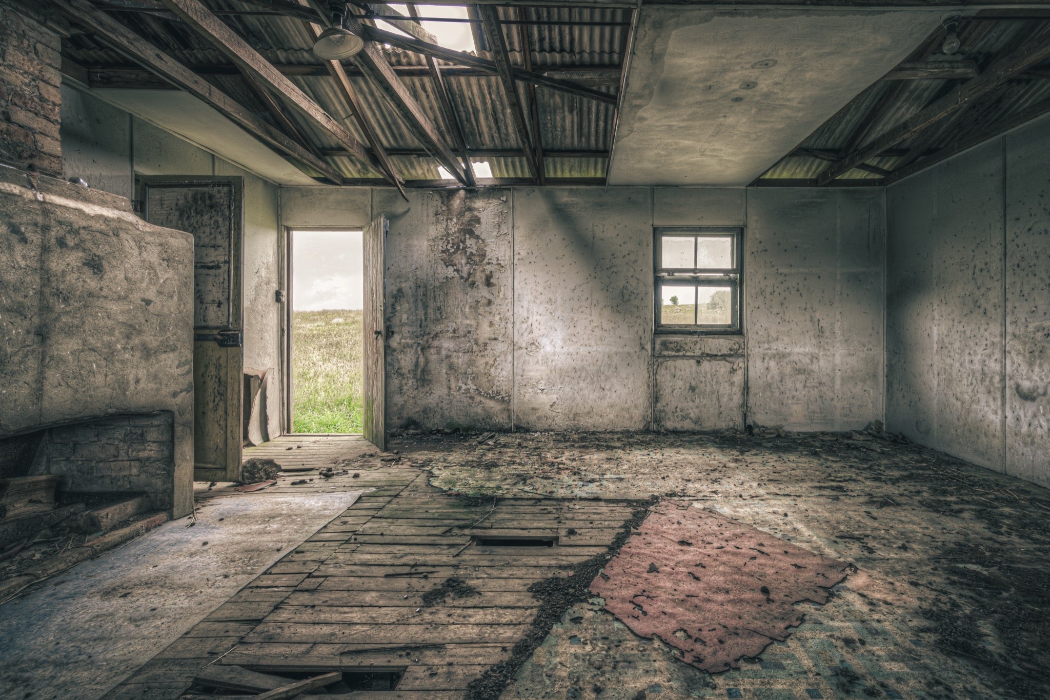 Interior of a derelict room at Jembaicumbene with a partly collapsed corrugated metal roof open to the sky, exposed timber rafters, rendered walls, a four-pane sash window, an open timber door to a paddock, and a torn red rug on broken floorboards.