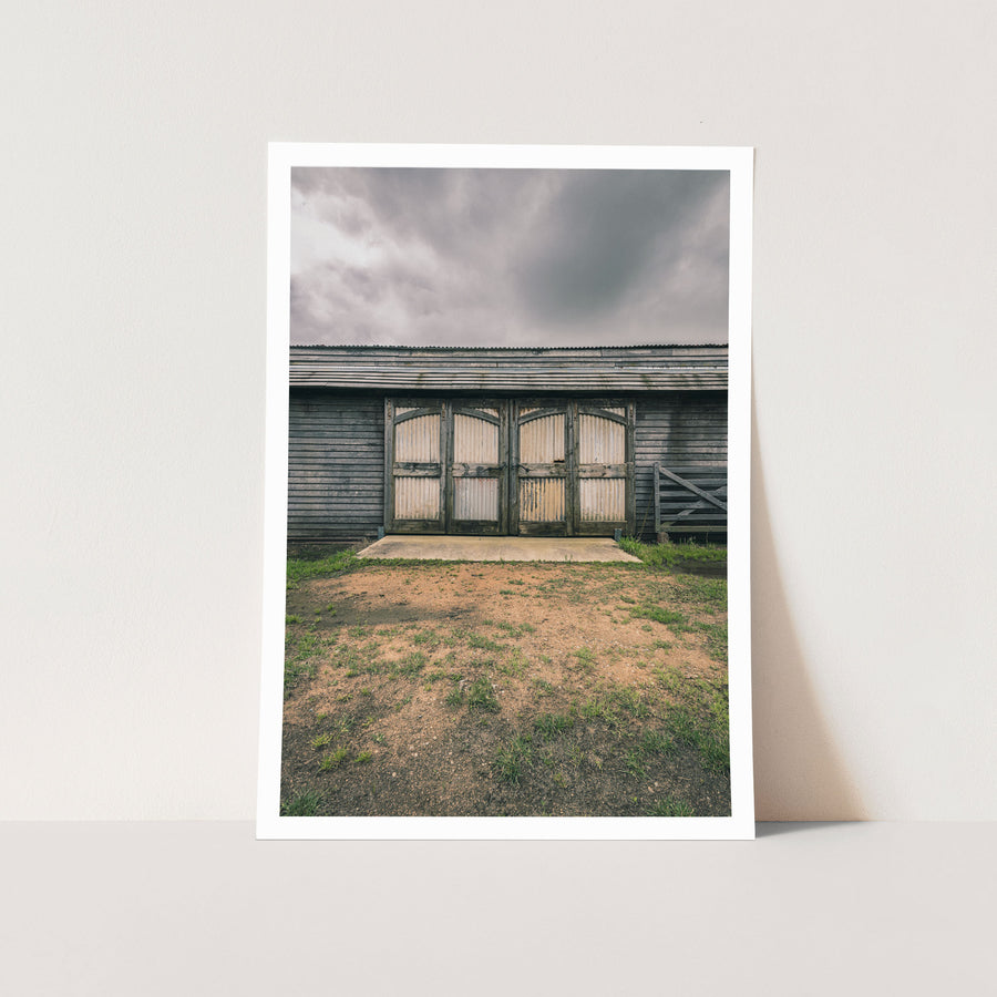 A pair of large corrugated metal barn doors with arched tops set into a weathered grey timber wall, with a concrete pad and timber farm gate to the right, at Jembaicumbene.