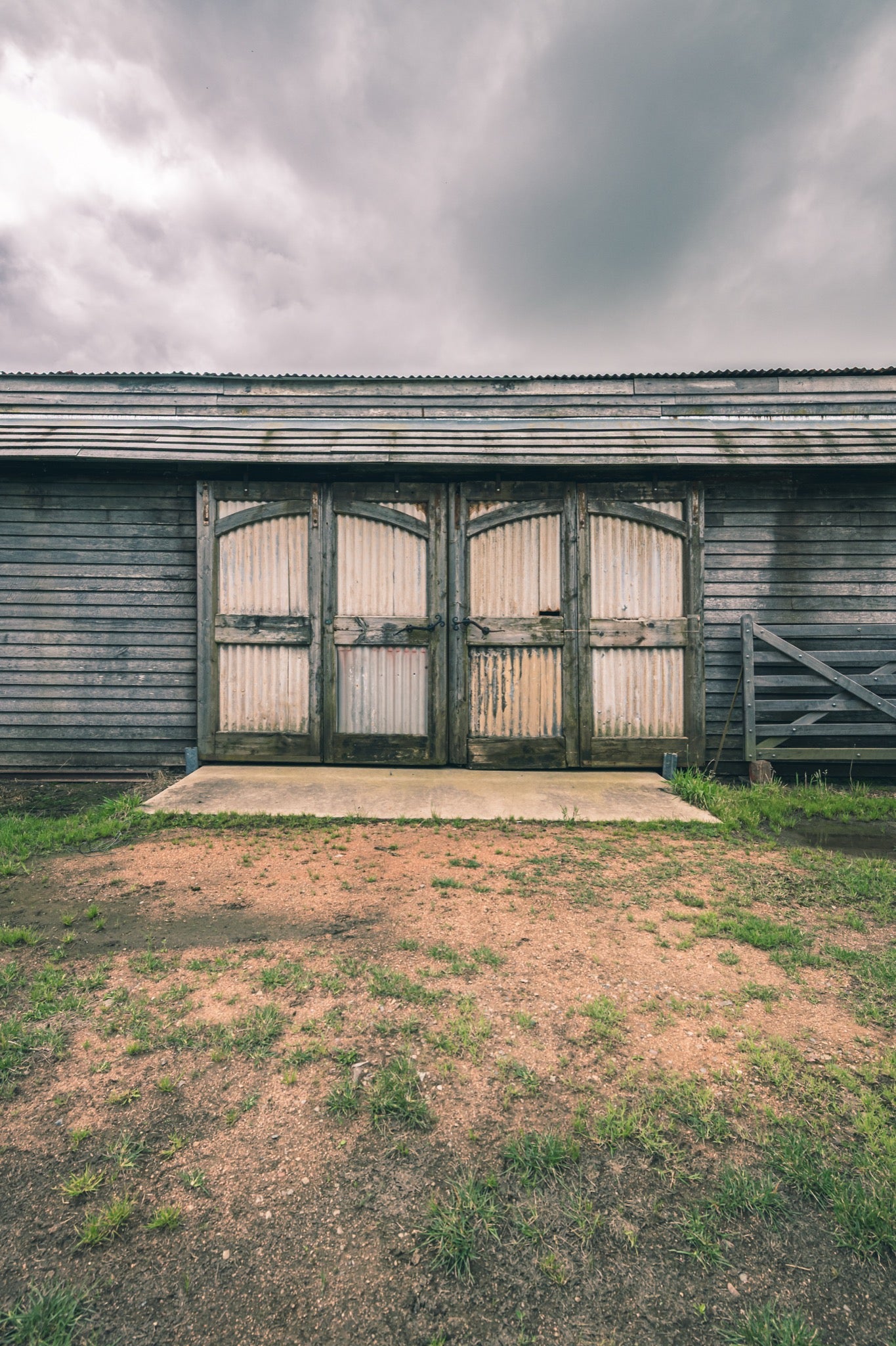 A pair of large corrugated metal barn doors with arched tops set into a weathered grey timber wall, with a concrete pad and timber farm gate to the right, at Jembaicumbene.