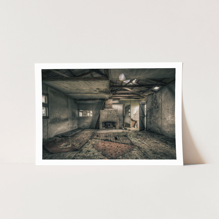 A rendered fireplace and brick chimney in a derelict room at Jembaicumbene, with whitewashed walls, a debris-strewn timber plank floor, and partly exposed roof framing overhead.