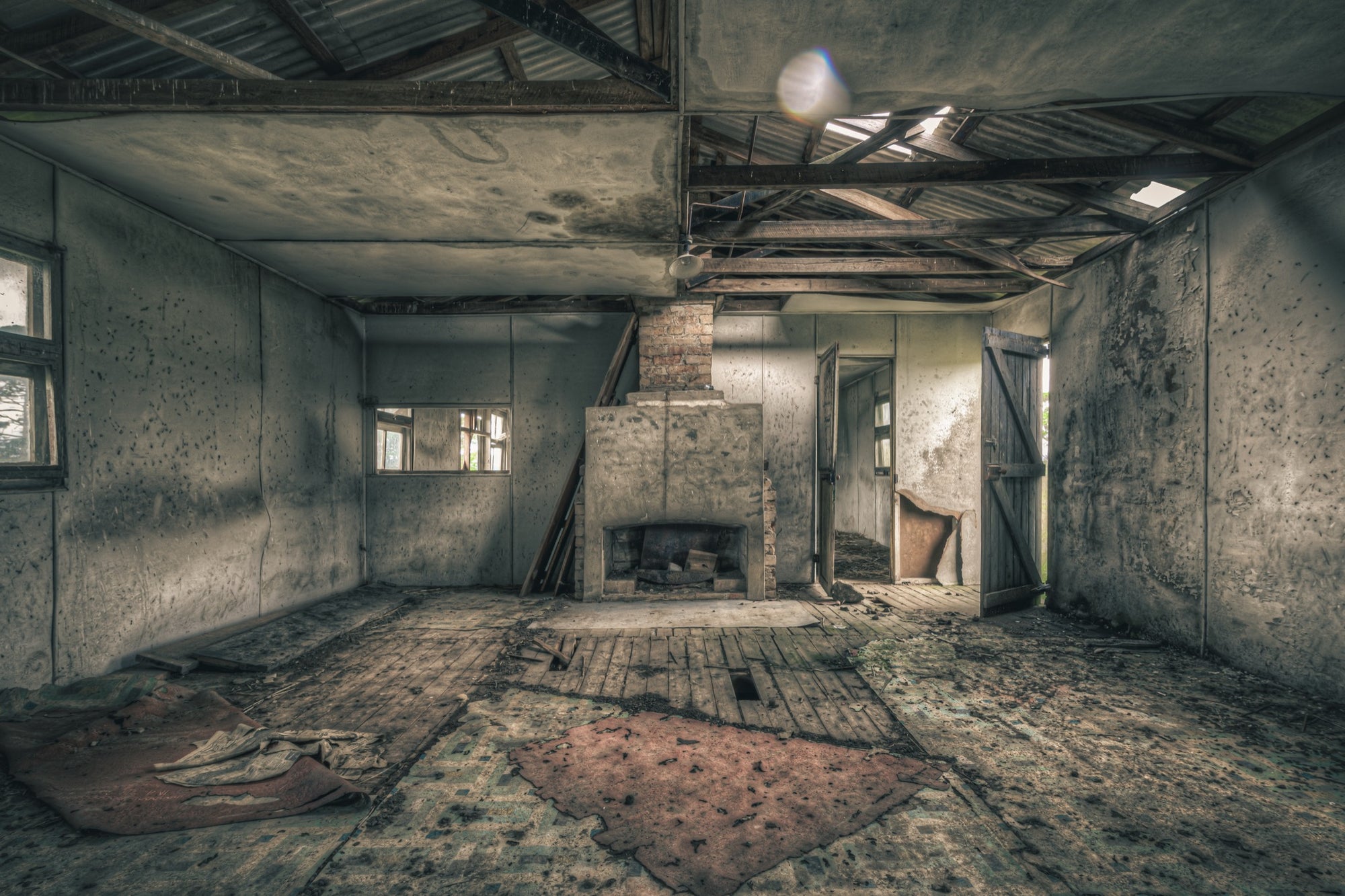 A rendered fireplace and brick chimney in a derelict room at Jembaicumbene, with whitewashed walls, a debris-strewn timber plank floor, and partly exposed roof framing overhead.