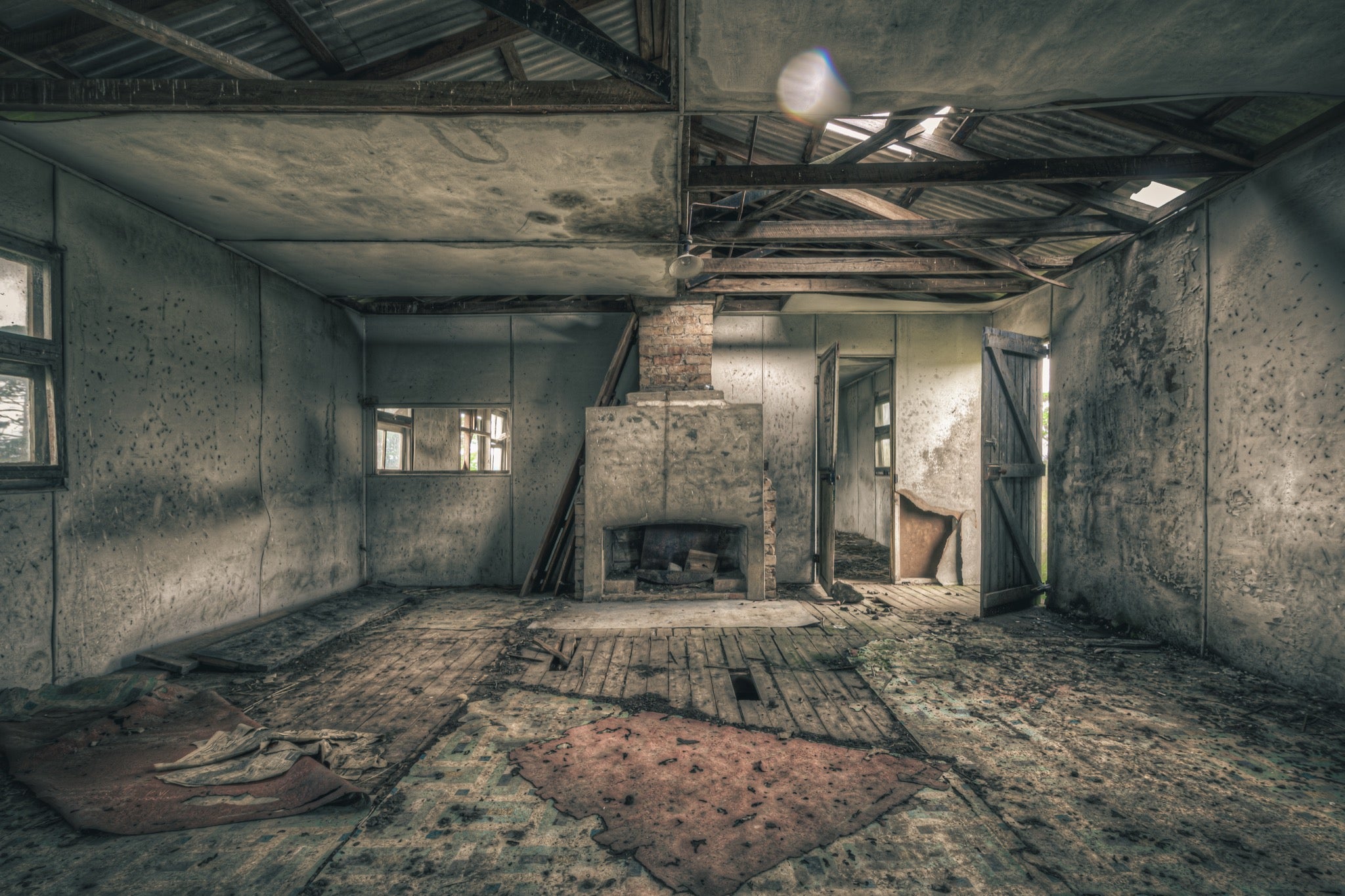 A rendered fireplace and brick chimney in a derelict room at Jembaicumbene, with whitewashed walls, a debris-strewn timber plank floor, and partly exposed roof framing overhead.