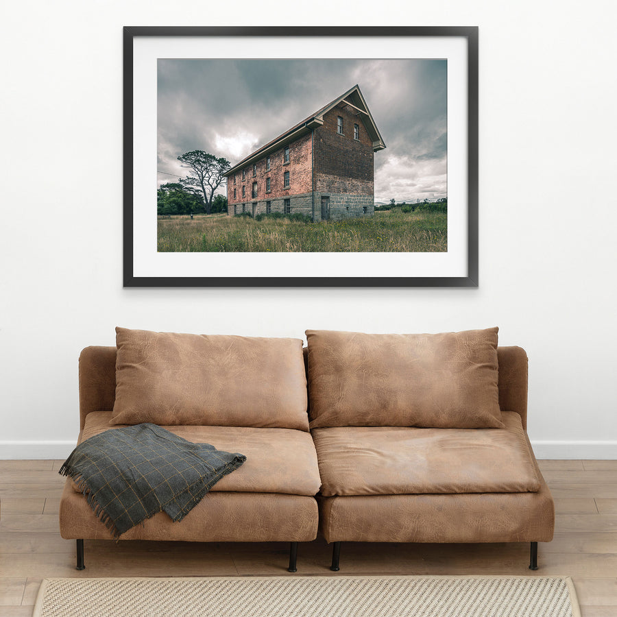The four-storey brick and stone flour mill at Jembaicumbene, its steep gabled end wall rising above long grass under a grey sky, with a timber door set into the granite base and small windows above.