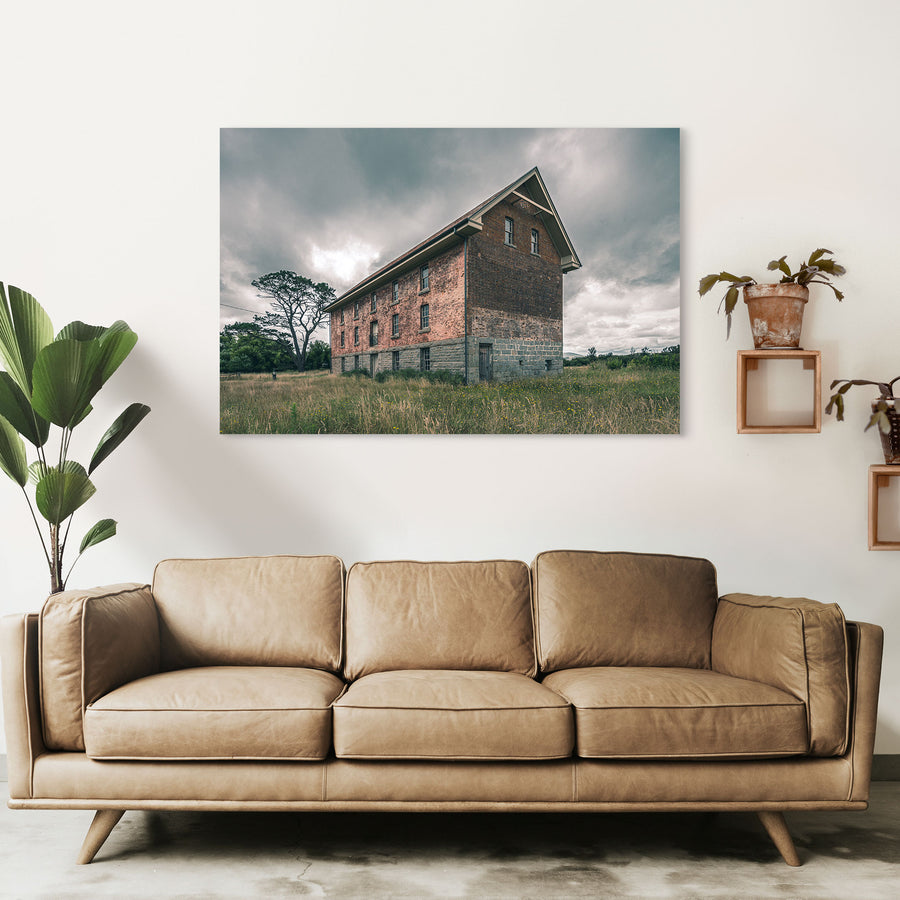 The four-storey brick and stone flour mill at Jembaicumbene, its steep gabled end wall rising above long grass under a grey sky, with a timber door set into the granite base and small windows above.