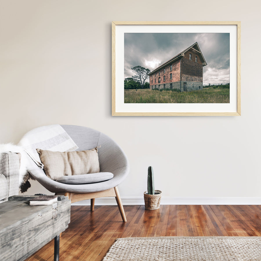 The four-storey brick and stone flour mill at Jembaicumbene, its steep gabled end wall rising above long grass under a grey sky, with a timber door set into the granite base and small windows above.