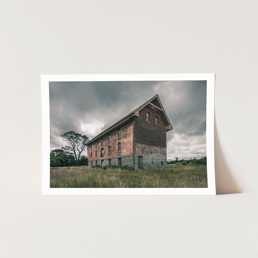 The four-storey brick and stone flour mill at Jembaicumbene, its steep gabled end wall rising above long grass under a grey sky, with a timber door set into the granite base and small windows above.