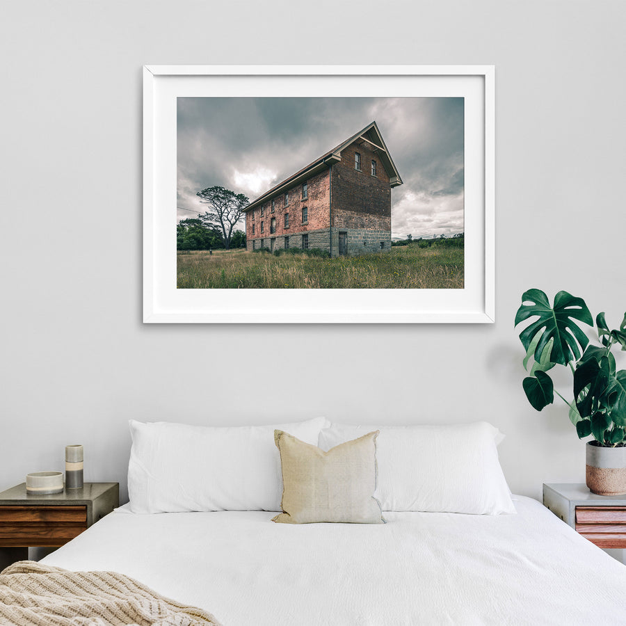 The four-storey brick and stone flour mill at Jembaicumbene, its steep gabled end wall rising above long grass under a grey sky, with a timber door set into the granite base and small windows above.