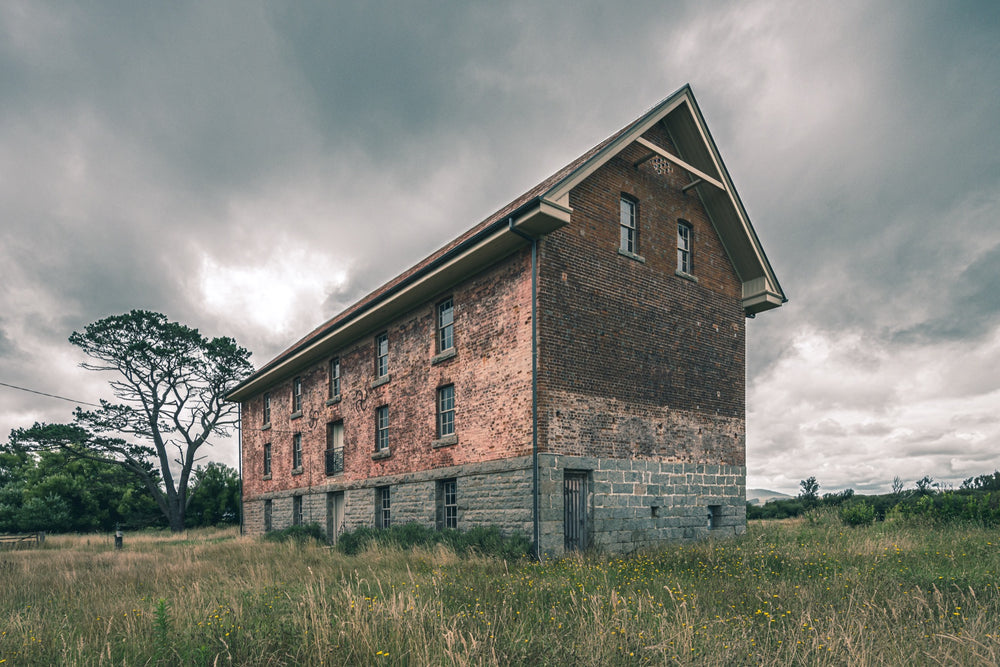 The four-storey brick and stone flour mill at Jembaicumbene, its steep gabled end wall rising above long grass under a grey sky, with a timber door set into the granite base and small windows above.