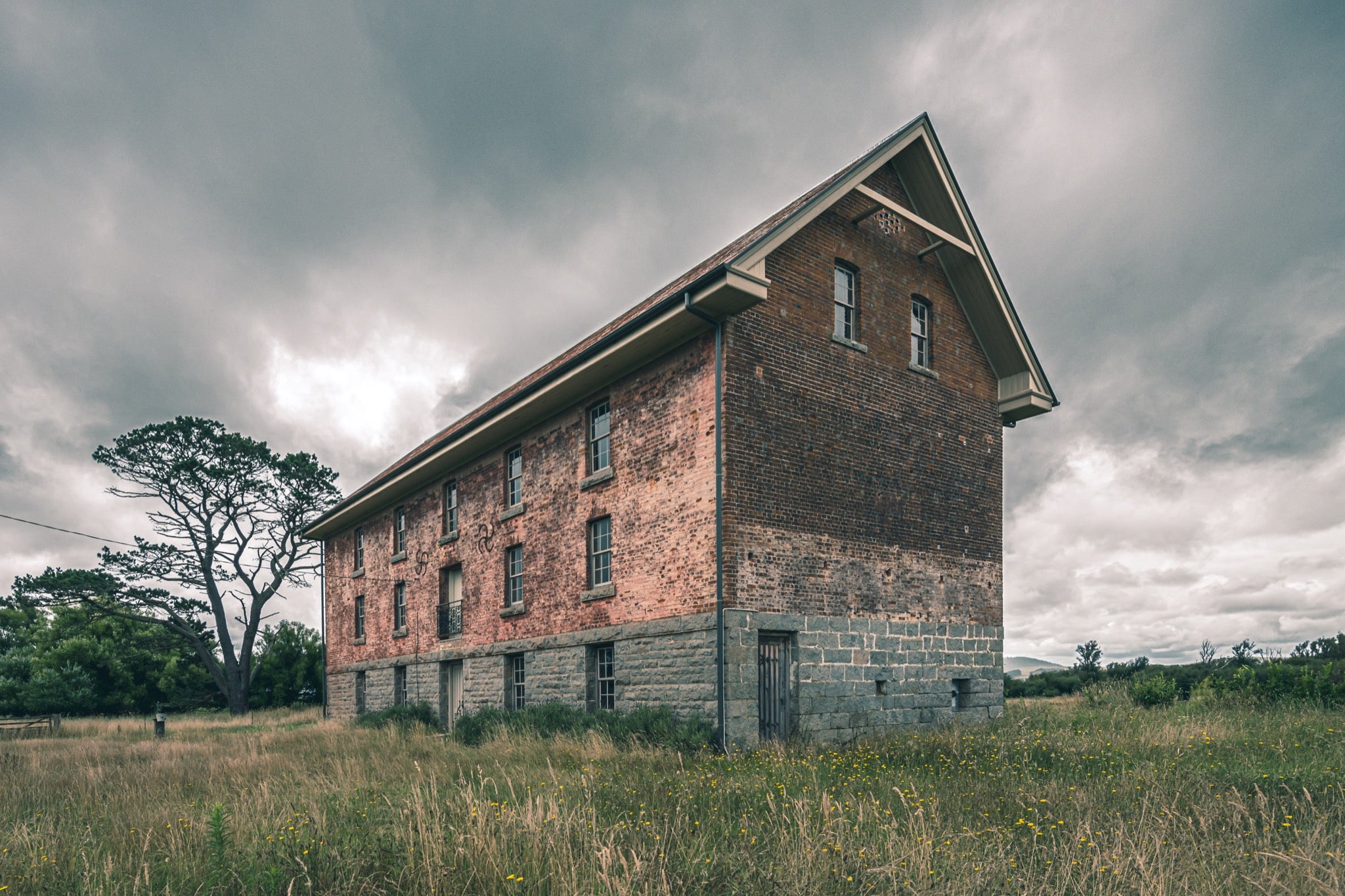 The four-storey brick and stone flour mill at Jembaicumbene, its steep gabled end wall rising above long grass under a grey sky, with a timber door set into the granite base and small windows above.