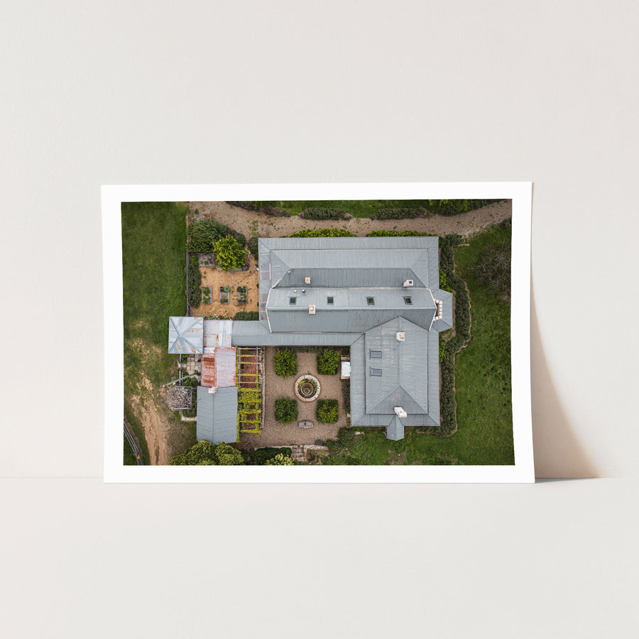 Aerial view of the formal gravel courtyard at Mill Pond Farm, Jembaicumbene, with a circular stone fountain at centre, clipped hedges, and grey corrugated metal roofs surrounding on three sides.