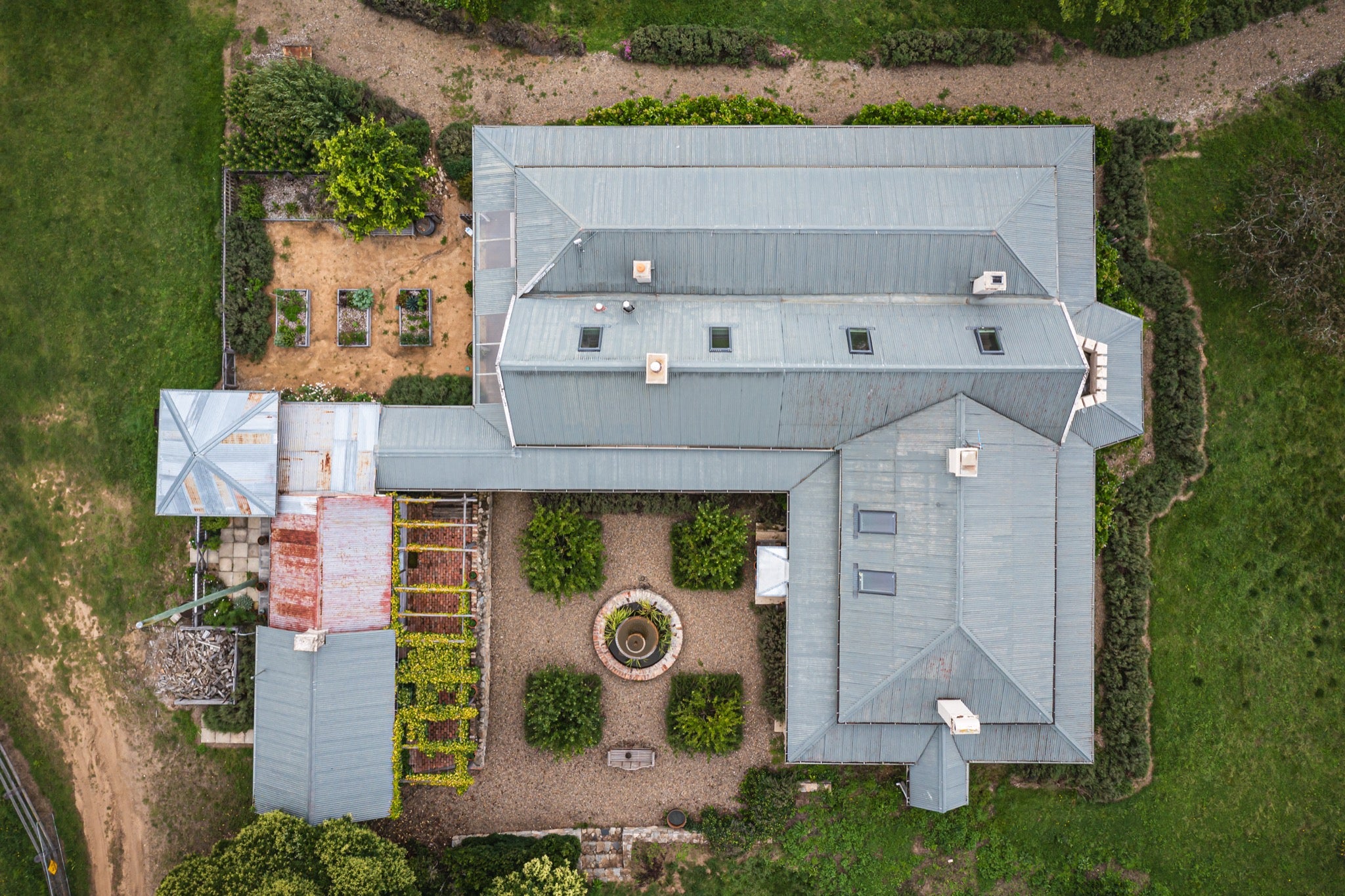 Aerial view of the formal gravel courtyard at Mill Pond Farm, Jembaicumbene, with a circular stone fountain at centre, clipped hedges, and grey corrugated metal roofs surrounding on three sides.