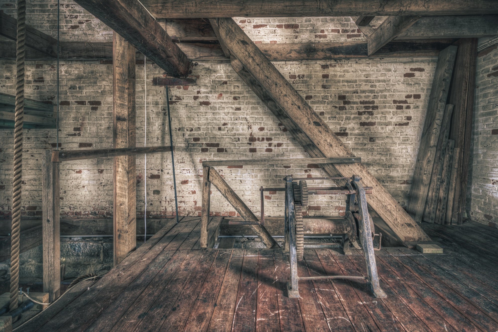 A cast iron gear and drum hoist mechanism on a heavy timber frame, with diagonal bracing beams and a whitewashed brick wall behind, photographed on an upper floor at Jembaicumbene.