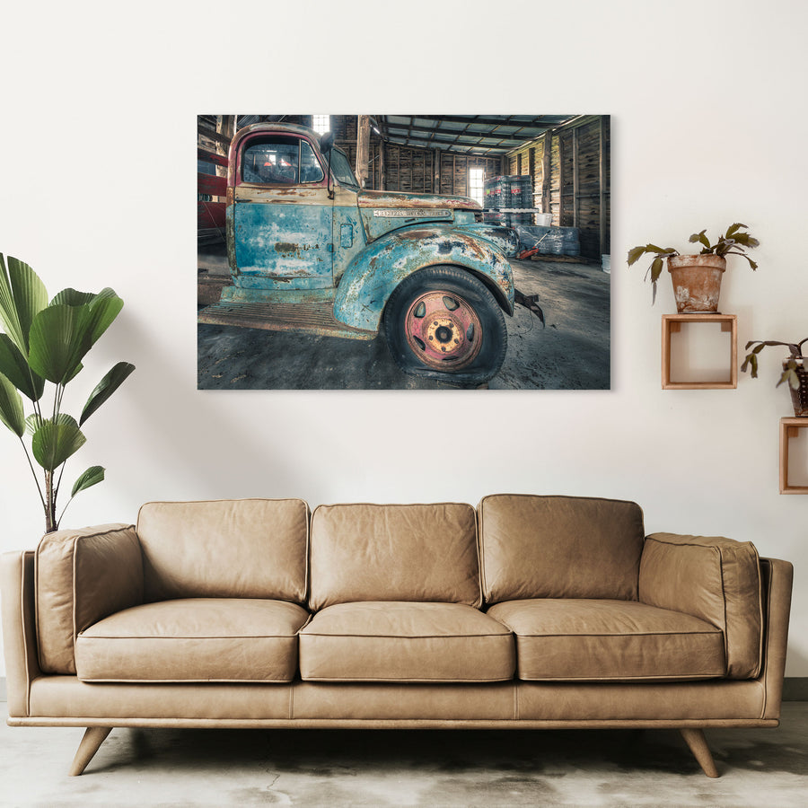 A rusting General Motors truck with faded blue paint and a pink and yellow steel wheel, parked inside a timber board barn at Jembaicumbene.