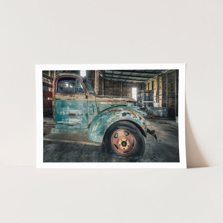 A rusting General Motors truck with faded blue paint and a pink and yellow steel wheel, parked inside a timber board barn at Jembaicumbene.