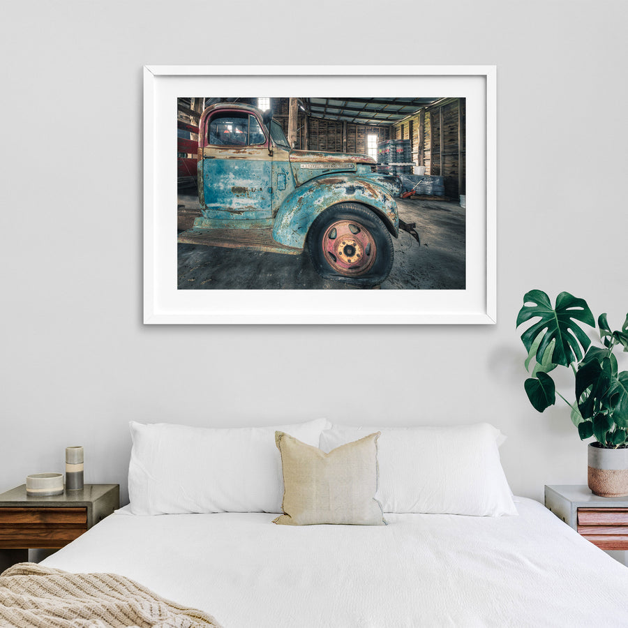 A rusting General Motors truck with faded blue paint and a pink and yellow steel wheel, parked inside a timber board barn at Jembaicumbene.