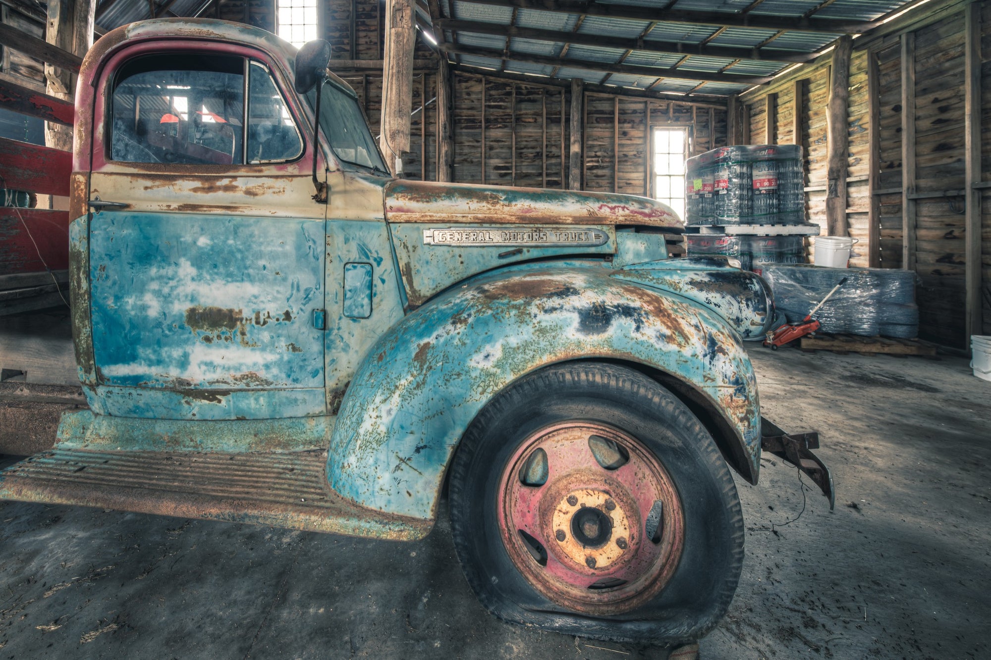 A rusting General Motors truck with faded blue paint and a pink and yellow steel wheel, parked inside a timber board barn at Jembaicumbene.