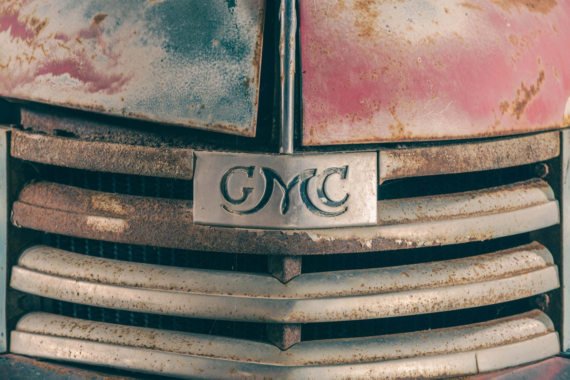 Chrome GMC nameplate centred on a rusted truck grille with worn horizontal bars and faded red and blue bodywork at Jembaicumbene.