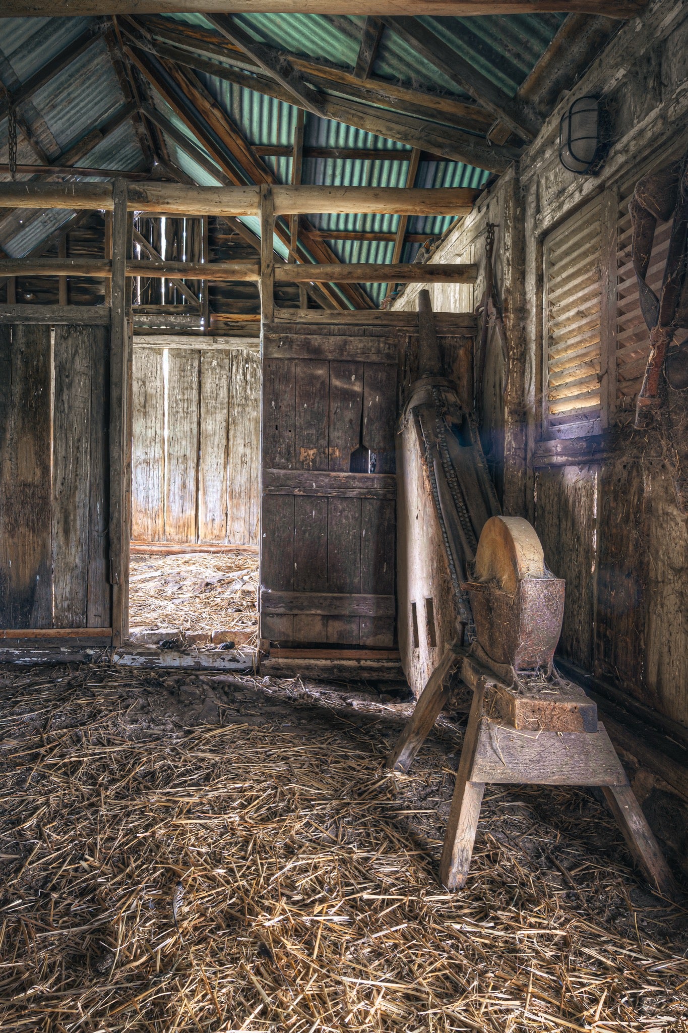A large stone wheel on a timber frame inside a weathered barn at Jembaicumbene, with a plank door, louvred window, straw-covered dirt floor, and corrugated iron roof on rough timber beams.