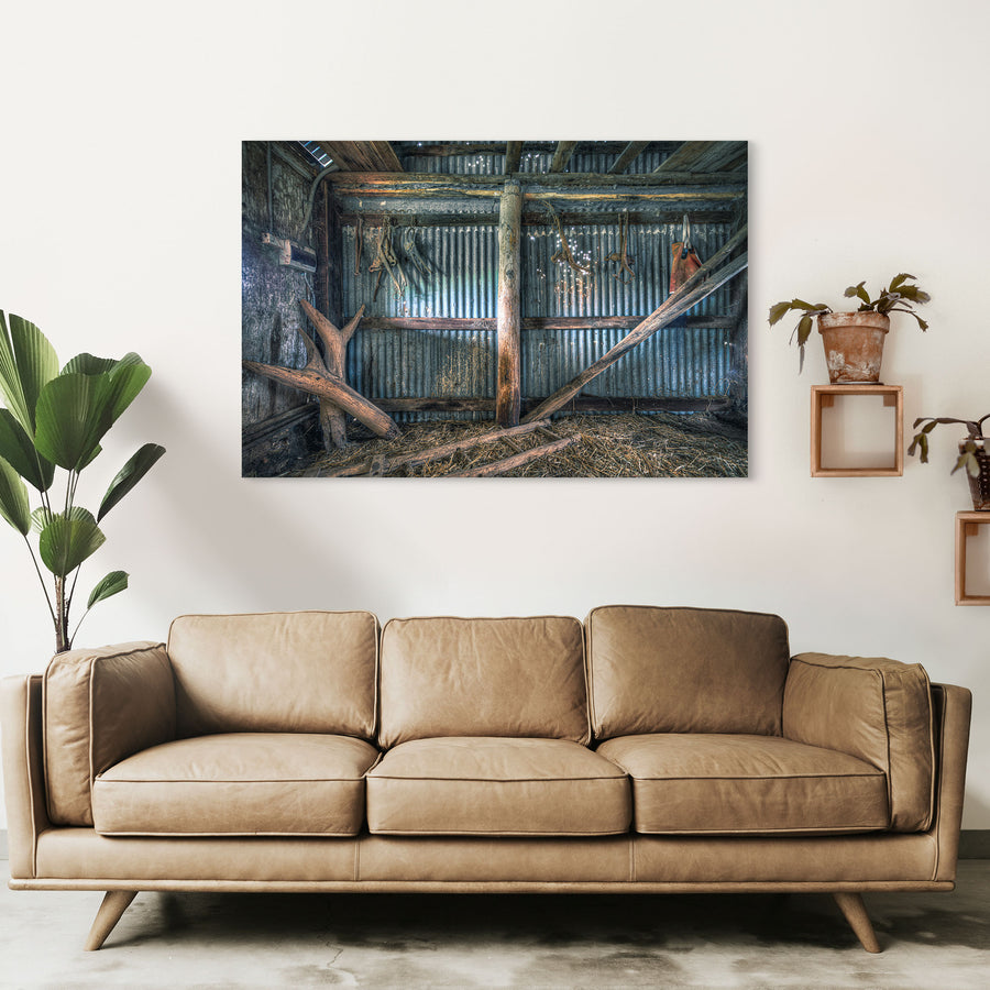 Old leather and metal harness pieces hang along a corrugated iron wall inside a timber shed at Jembaicumbene, with fallen beams and loose straw covering the dirt floor.