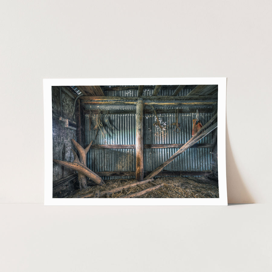 Old leather and metal harness pieces hang along a corrugated iron wall inside a timber shed at Jembaicumbene, with fallen beams and loose straw covering the dirt floor.