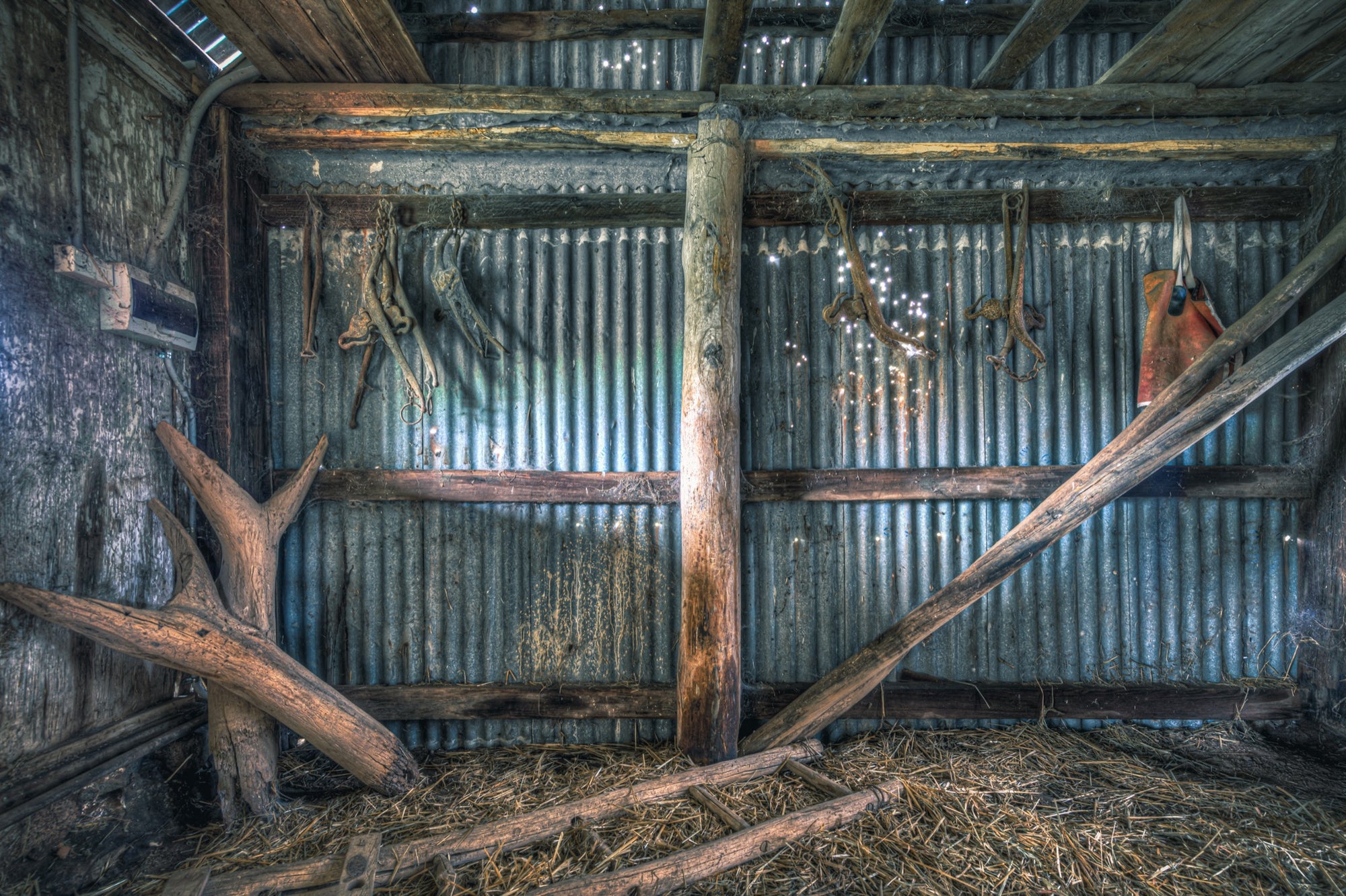 Old leather and metal harness pieces hang along a corrugated iron wall inside a timber shed at Jembaicumbene, with fallen beams and loose straw covering the dirt floor.