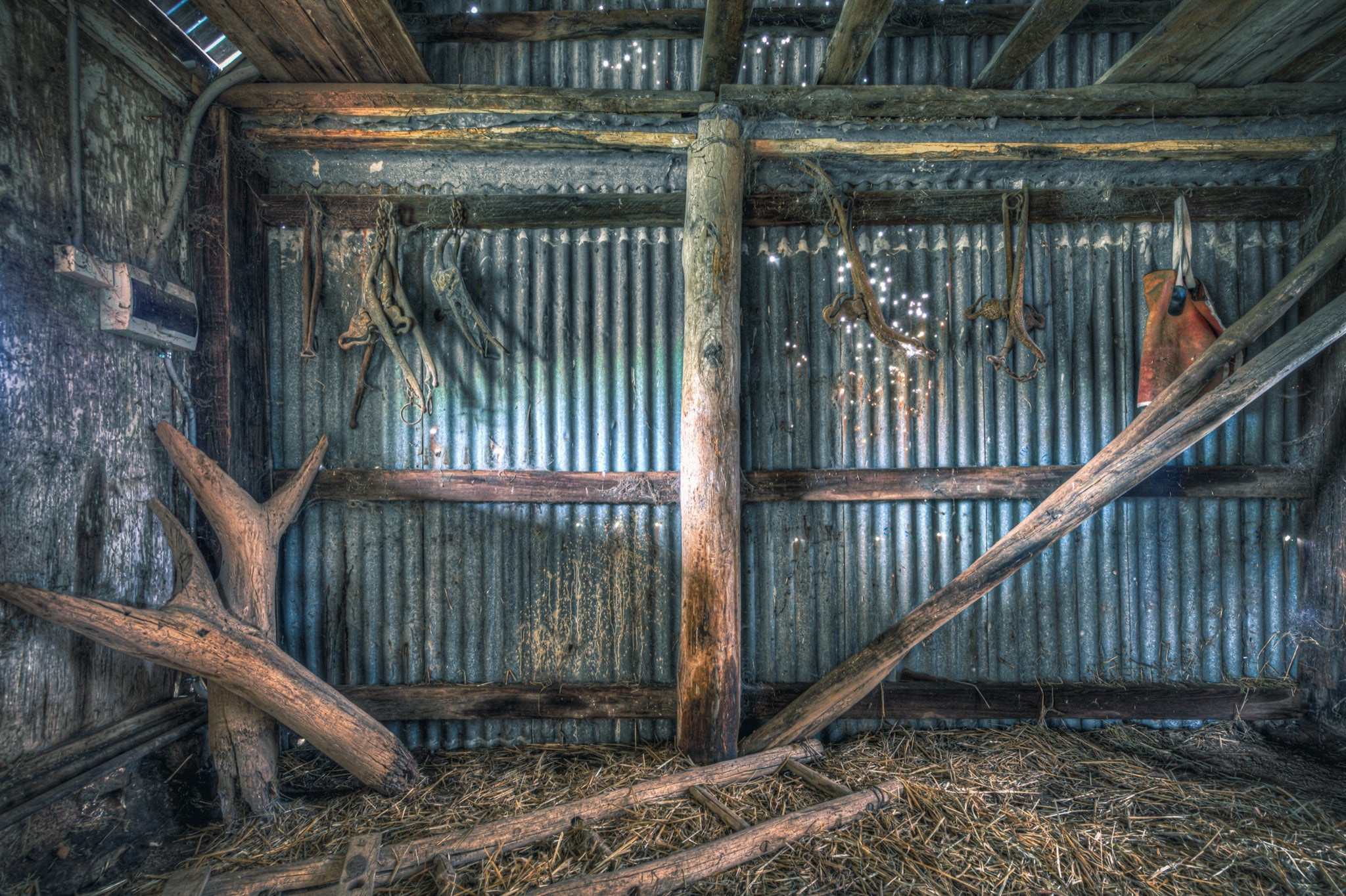Old leather and metal harness pieces hang along a corrugated iron wall inside a timber shed at Jembaicumbene, with fallen beams and loose straw covering the dirt floor.