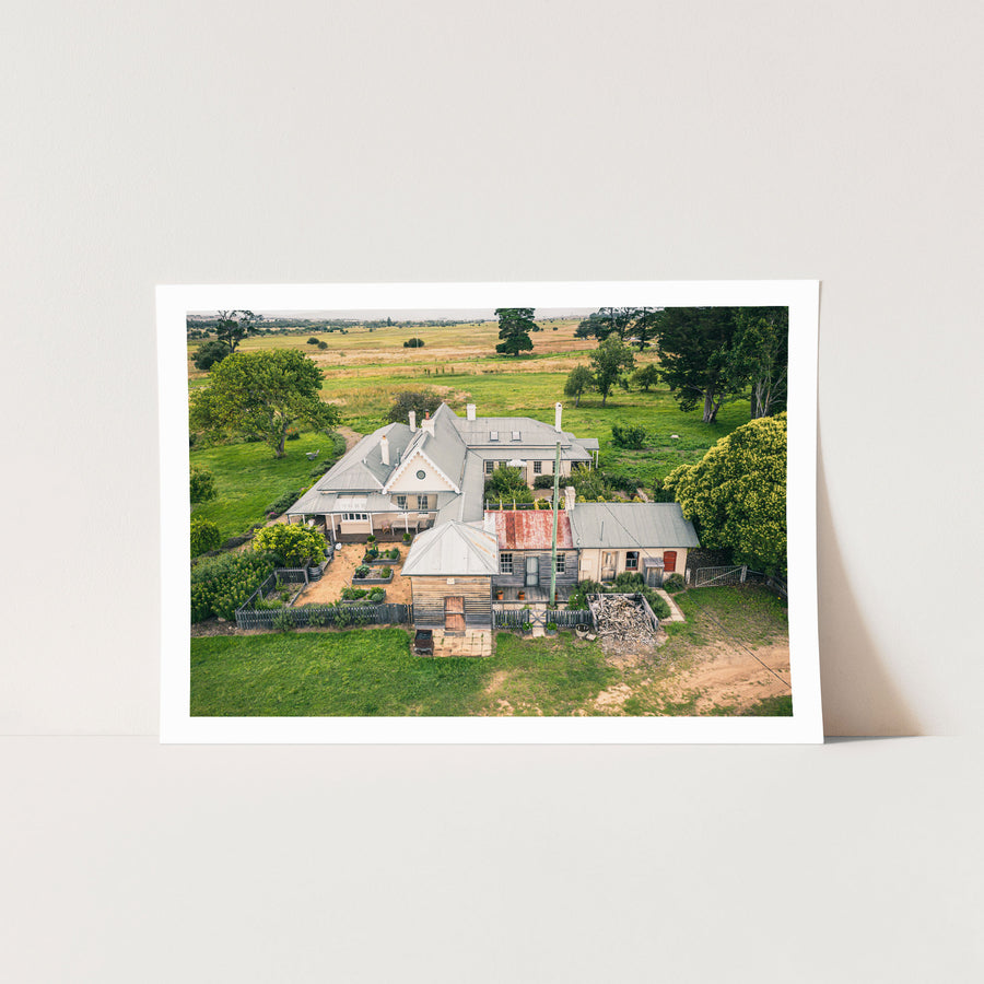 Aerial view of the Mill Pond Farm homestead at Jembaicumbene, showing hipped grey roofs, chimneys, a corrugated iron outbuilding, a timber barn, a vegetable garden, and surrounding green paddocks.