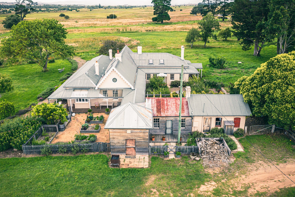 Aerial view of the Mill Pond Farm homestead at Jembaicumbene, showing hipped grey roofs, chimneys, a corrugated iron outbuilding, a timber barn, a vegetable garden, and surrounding green paddocks.